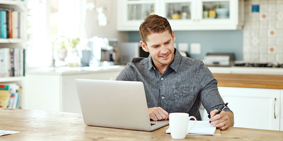 Mann sitter på kjøkkenet og skriver i notatblokk med en kaffekopp ved siden og PC fremfor seg, til illustrasjon for hvordan beregne egenkapital