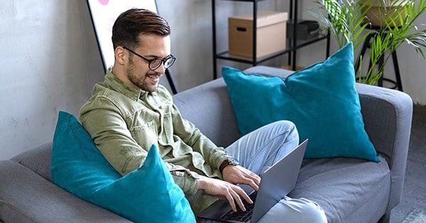 Mann sitter i grå sofa med lyseblå puter og jobber på PC, til illustrasjon for sett opp budsjett