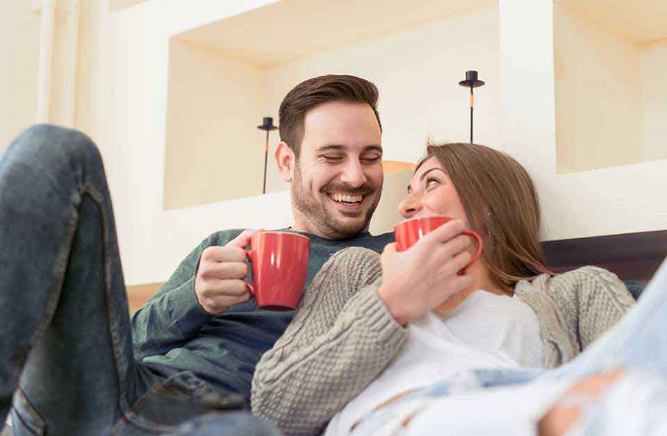 Mann med kaffekopp og kvinne som lener seg på mannen med kaffekopp smiler i sofa, til illustrasjon for hvordan få pengene til å vare