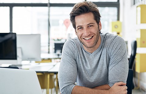 Mann sitter ved kontorplass med datamaskin fremfor seg og smiler, til illustrasjon for spareavtale