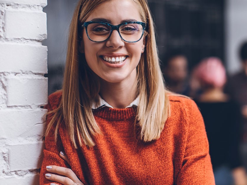 Kvinne med orange genser og briller smiler i kontorlokale, til illustrasjon for bli bedriftskunde