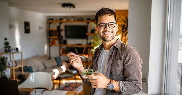 Mann står i stua og smiler mens han spiser en salat, til illustrasjon for refinansieringslån