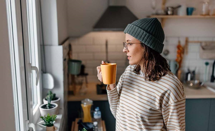 Dame med lue, briller og stripete genser står på kjøkkenet og drikker kaffe